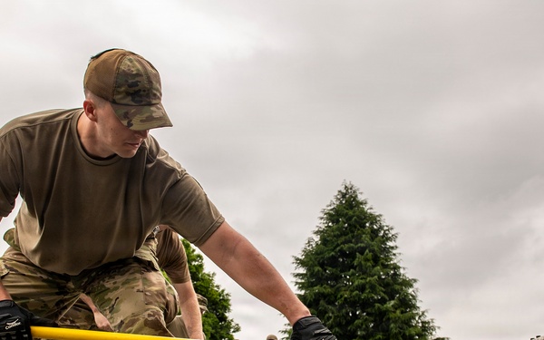 Military personnel in MultiCam/OCP camouflage uniforms are working to level or screed freshly poured concrete using a yellow screed bar. The foreground soldier is leaning forward with both gloved hands gripping the tool, actively smoothing the wet concrete surface. A metal rebar/reinforcement framework is visible embedded in the concrete, and additional personnel and construction equipment are visible in the background.