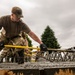 Military personnel in MultiCam/OCP camouflage uniforms are working to level or screed freshly poured concrete using a yellow screed bar. The foreground soldier is leaning forward with both gloved hands gripping the tool, actively smoothing the wet concrete surface. A metal rebar/reinforcement framework is visible embedded in the concrete, and additional personnel and construction equipment are visible in the background.