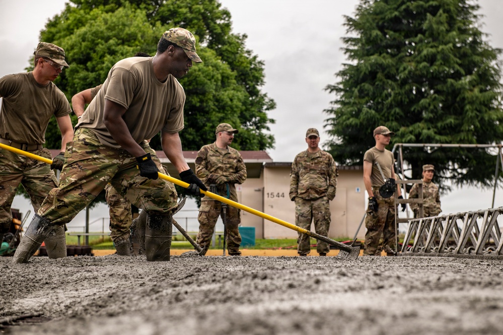 Military personnel in OCP (Operational Camouflage Pattern) uniforms are working together to spread and level freshly poured concrete on a surface. Two soldiers in the foreground are actively using yellow-handled rakes/lutes to distribute the wet concrete, while several others observe in the background. The setting appears to be a military installation, indicated by a building numbered '1544' visible in the background.
