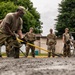 Military personnel in OCP (Operational Camouflage Pattern) uniforms are working together to spread and level freshly poured concrete on a surface. Two soldiers in the foreground are actively using yellow-handled rakes/lutes to distribute the wet concrete, while several others observe in the background. The setting appears to be a military installation, indicated by a building numbered '1544' visible in the background.