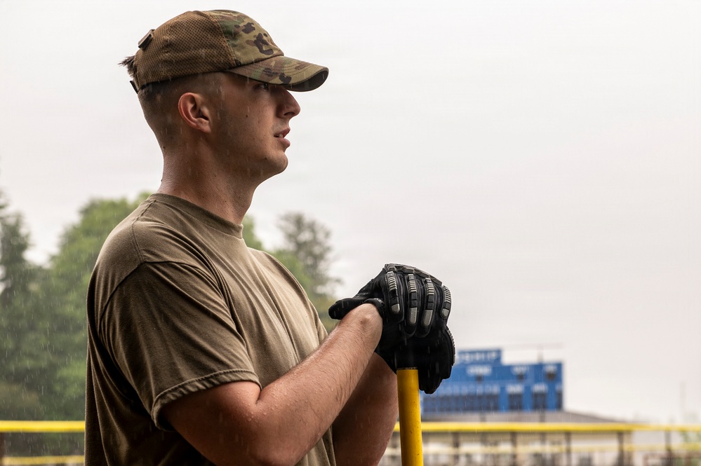 A young male service member in military-style clothing stands outdoors in light rain, holding a yellow-handled tool with both gloved hands. He is wearing a multicam/OCP pattern cap and an olive drab t-shirt, gazing into the distance. The background suggests an outdoor work or community service environment near a sports facility.