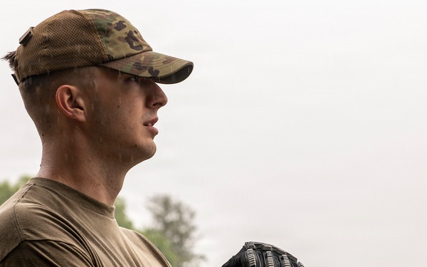 A young male service member in military-style clothing stands outdoors in light rain, holding a yellow-handled tool with both gloved hands. He is wearing a multicam/OCP pattern cap and an olive drab t-shirt, gazing into the distance. The background suggests an outdoor work or community service environment near a sports facility.