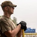 A young male service member in military-style clothing stands outdoors in light rain, holding a yellow-handled tool with both gloved hands. He is wearing a multicam/OCP pattern cap and an olive drab t-shirt, gazing into the distance. The background suggests an outdoor work or community service environment near a sports facility.