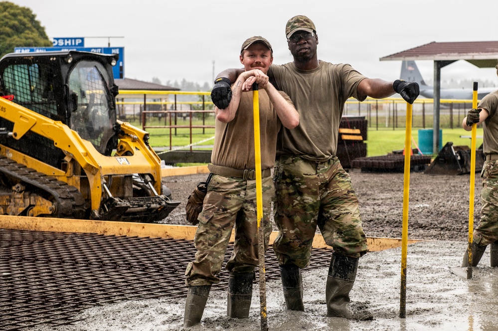 Two military personnel in OCP (Operational Camouflage Pattern) uniforms pose for the camera while working in muddy conditions at what appears to be a military base construction or renovation site. They are wearing rubber boots and tactical gloves, leaning on yellow-handled tools. A Caterpillar skid steer loader is visible in the background along with what appears to be a military aircraft on a flight line.