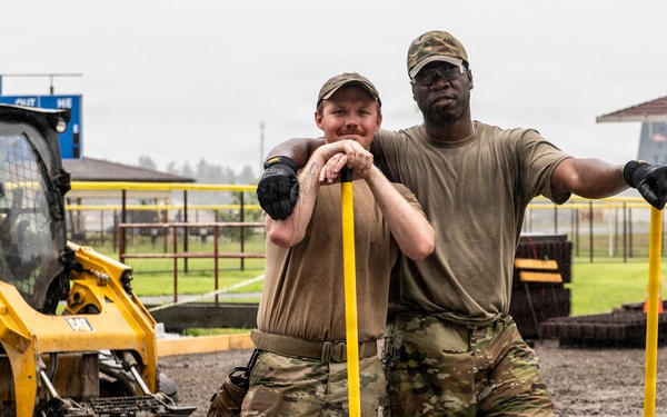 Two military personnel in OCP (Operational Camouflage Pattern) uniforms pose for the camera while working in muddy conditions at what appears to be a military base construction or renovation site. They are wearing rubber boots and tactical gloves, leaning on yellow-handled tools. A Caterpillar skid steer loader is visible in the background along with what appears to be a military aircraft on a flight line.