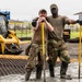 Two military personnel in OCP (Operational Camouflage Pattern) uniforms pose for the camera while working in muddy conditions at what appears to be a military base construction or renovation site. They are wearing rubber boots and tactical gloves, leaning on yellow-handled tools. A Caterpillar skid steer loader is visible in the background along with what appears to be a military aircraft on a flight line.