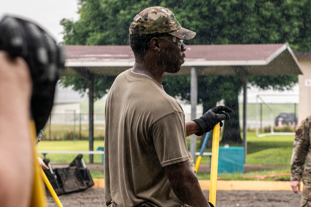 A military service member wearing a multicam cap, olive drab t-shirt, and black work gloves is working outdoors in the rain, holding a yellow-handled tool. The scene appears to be a construction or maintenance work detail on what looks like a military installation. At least one other uniformed individual is visible in the background.