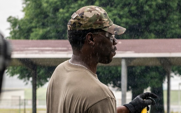 A military service member wearing a multicam cap, olive drab t-shirt, and black work gloves is working outdoors in the rain, holding a yellow-handled tool. The scene appears to be a construction or maintenance work detail on what looks like a military installation. At least one other uniformed individual is visible in the background.