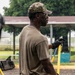 A military service member wearing a multicam cap, olive drab t-shirt, and black work gloves is working outdoors in the rain, holding a yellow-handled tool. The scene appears to be a construction or maintenance work detail on what looks like a military installation. At least one other uniformed individual is visible in the background.