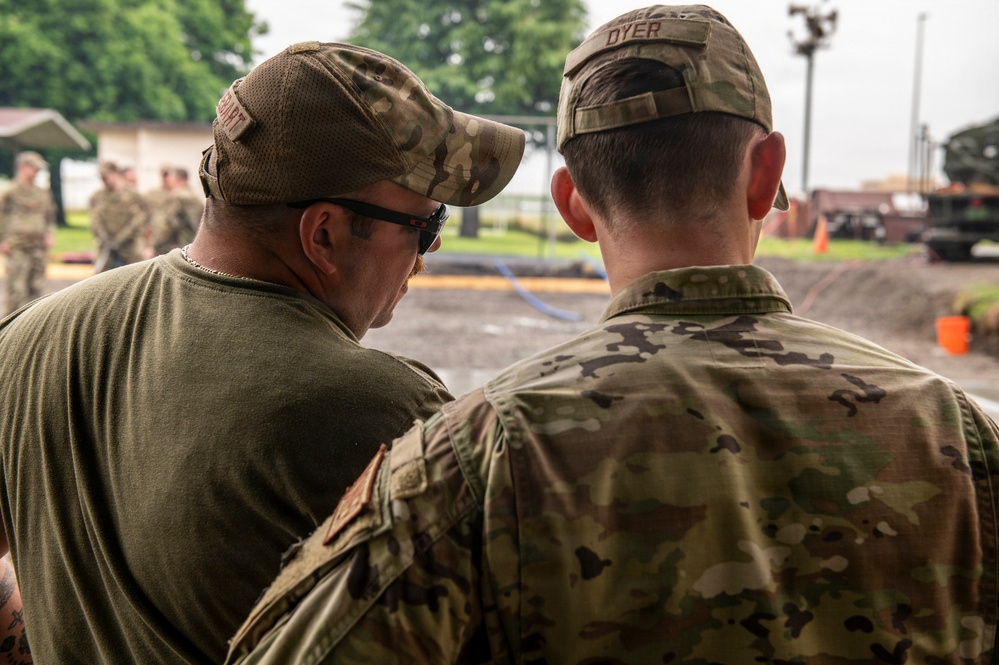Two military personnel are seen from behind, appearing to confer or observe an activity in the foreground. The individual on the right wears a full OCP (Operational Camouflage Pattern) uniform with a name tape reading 'DYER', while the individual on the left wears a green t-shirt with a camouflage cap and sunglasses. In the background, additional uniformed personnel and military vehicles are visible in what appears to be a training or operational area.