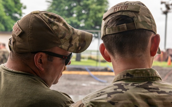 Two military personnel are seen from behind, appearing to confer or observe an activity in the foreground. The individual on the right wears a full OCP (Operational Camouflage Pattern) uniform with a name tape reading 'DYER', while the individual on the left wears a green t-shirt with a camouflage cap and sunglasses. In the background, additional uniformed personnel and military vehicles are visible in what appears to be a training or operational area.