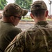 Two military personnel are seen from behind, appearing to confer or observe an activity in the foreground. The individual on the right wears a full OCP (Operational Camouflage Pattern) uniform with a name tape reading 'DYER', while the individual on the left wears a green t-shirt with a camouflage cap and sunglasses. In the background, additional uniformed personnel and military vehicles are visible in what appears to be a training or operational area.