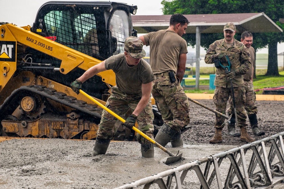Military personnel in camouflage uniforms are working in heavy rain, shoveling and spreading material in a muddy, waterlogged area. A yellow CAT skid-steer loader (max 2,000 lbs capacity) is positioned nearby, likely having deposited gravel or fill material. The scene appears to be a construction or flood-response operation at a military installation.