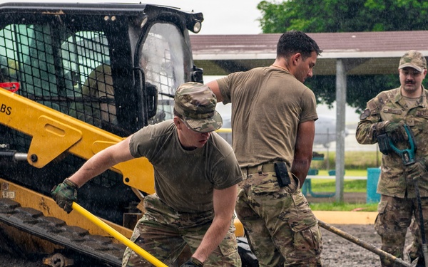 Military personnel in camouflage uniforms are working in heavy rain, shoveling and spreading material in a muddy, waterlogged area. A yellow CAT skid-steer loader (max 2,000 lbs capacity) is positioned nearby, likely having deposited gravel or fill material. The scene appears to be a construction or flood-response operation at a military installation.
