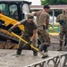Military personnel in camouflage uniforms are working in heavy rain, shoveling and spreading material in a muddy, waterlogged area. A yellow CAT skid-steer loader (max 2,000 lbs capacity) is positioned nearby, likely having deposited gravel or fill material. The scene appears to be a construction or flood-response operation at a military installation.