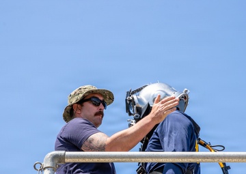 Two individuals are on an elevated platform, with one person assisting the other in donning or adjusting a diving helmet. The scene appears to be a military or naval diving operation, with specialized diving equipment visible including a hard-hat diving helmet and associated gear. The setting is outdoors under a clear blue sky, likely aboard a ship or dock.
