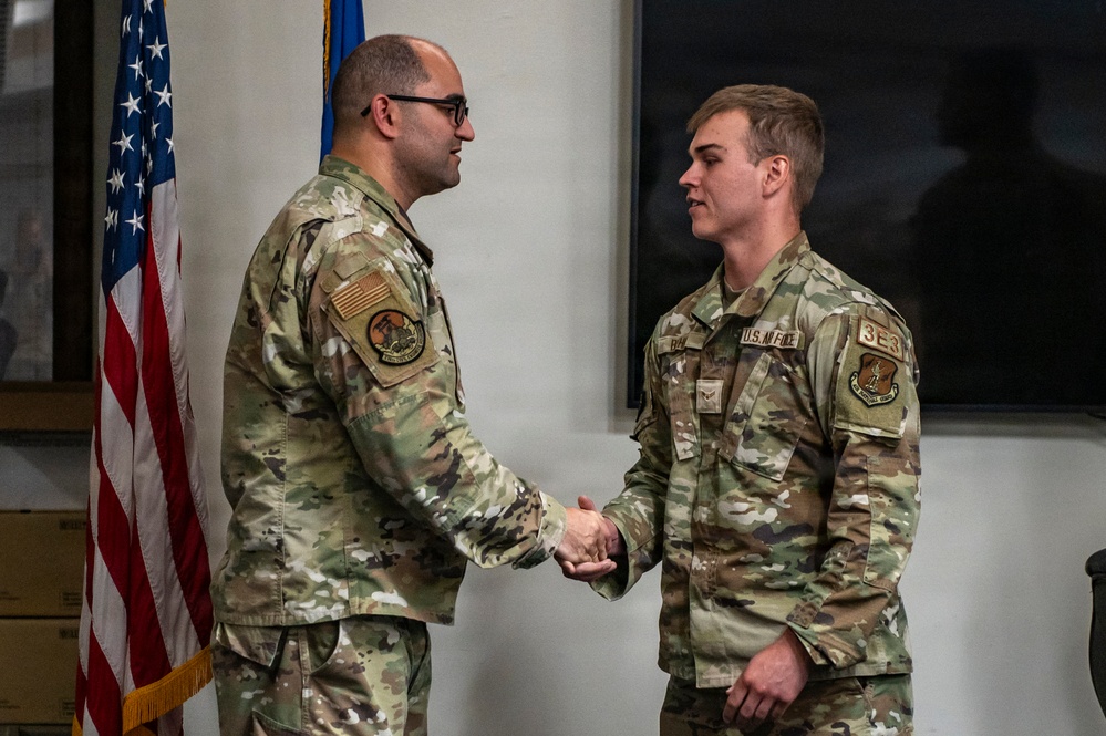 Two U.S. Air Force personnel in Operational Camouflage Pattern (OCP) uniforms are shaking hands in what appears to be a formal military ceremony or recognition event. The individual on the right has a visible '3E3' identifier patch and 'U.S. AIR FORCE' tape on their uniform. An American flag and what appears to be a unit flag are visible in the background.