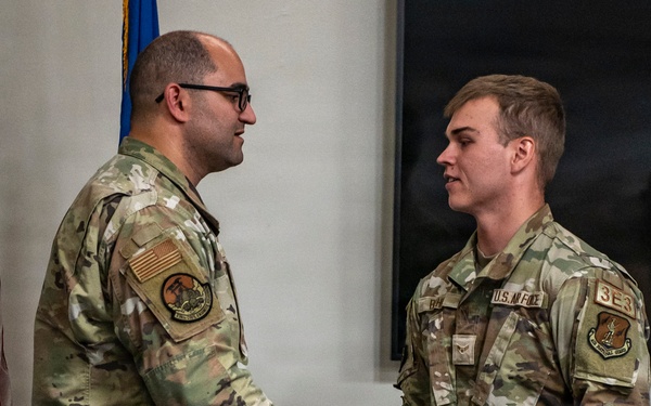 Two U.S. Air Force personnel in Operational Camouflage Pattern (OCP) uniforms are shaking hands in what appears to be a formal military ceremony or recognition event. The individual on the right has a visible '3E3' identifier patch and 'U.S. AIR FORCE' tape on their uniform. An American flag and what appears to be a unit flag are visible in the background.
