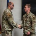 Two U.S. Air Force personnel in Operational Camouflage Pattern (OCP) uniforms are shaking hands in what appears to be a formal military ceremony or recognition event. The individual on the right has a visible '3E3' identifier patch and 'U.S. AIR FORCE' tape on their uniform. An American flag and what appears to be a unit flag are visible in the background.