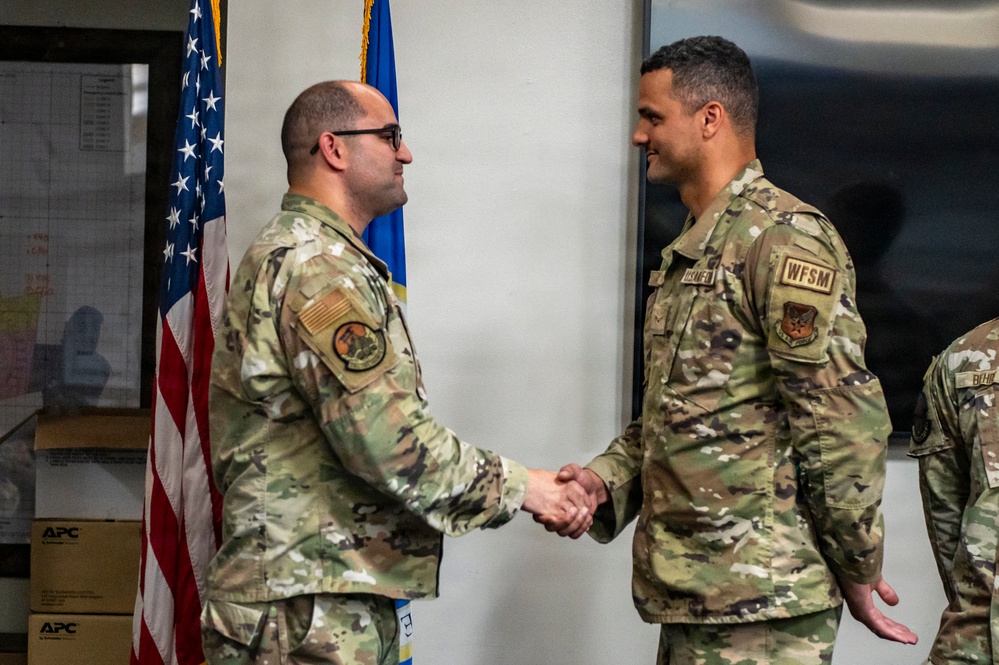 Two U.S. Air Force personnel in OCP (Operational Camouflage Pattern) uniforms are shaking hands in what appears to be an official ceremony or recognition event. An American flag and an Air Force flag are visible in the background. A third uniformed individual is partially visible on the right edge of the frame.