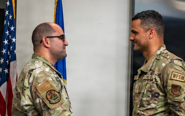 Two U.S. Air Force personnel in OCP (Operational Camouflage Pattern) uniforms are shaking hands in what appears to be an official ceremony or recognition event. An American flag and an Air Force flag are visible in the background. A third uniformed individual is partially visible on the right edge of the frame.
