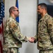 Two U.S. Air Force personnel in OCP (Operational Camouflage Pattern) uniforms are shaking hands in what appears to be an official ceremony or recognition event. An American flag and an Air Force flag are visible in the background. A third uniformed individual is partially visible on the right edge of the frame.