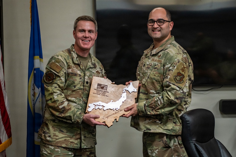 Two U.S. Air Force personnel in OCP (Operational Camouflage Pattern) uniforms are posing together while holding a wooden plaque shaped like Japan. The plaque appears to be a commemorative award or recognition item with an engraved map of Japan and text including 'Engineers lead the way.' The setting appears to be an official military office with U.S. Air Force flags visible in the background.