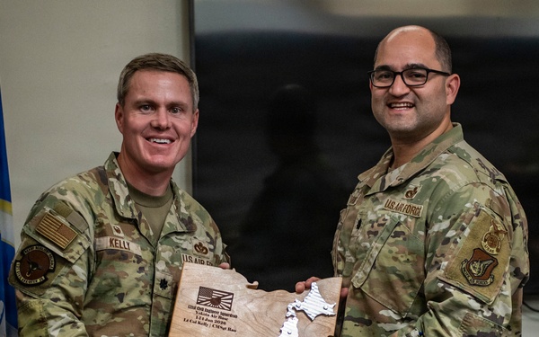 Two U.S. Air Force personnel in OCP (Operational Camouflage Pattern) uniforms are posing together while holding a wooden plaque shaped like Japan. The plaque appears to be a commemorative award or recognition item with an engraved map of Japan and text including 'Engineers lead the way.' The setting appears to be an official military office with U.S. Air Force flags visible in the background.