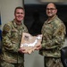 Two U.S. Air Force personnel in OCP (Operational Camouflage Pattern) uniforms are posing together while holding a wooden plaque shaped like Japan. The plaque appears to be a commemorative award or recognition item with an engraved map of Japan and text including 'Engineers lead the way.' The setting appears to be an official military office with U.S. Air Force flags visible in the background.