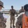 A group of military personnel in desert camouflage uniforms are gathered outdoors in an arid environment, appearing to conduct a briefing or training exercise. A central figure is speaking and gesturing while holding documents, with several others listening, some wearing orange high-visibility vests marked 'IC'. The setting appears to be a military installation in a desert region.