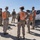 A group of U.S. Marine Corps personnel in desert camouflage uniforms are gathered in an arid outdoor environment, likely during a training exercise. Several individuals are wearing bright orange identification vests/backpacks with alphanumeric labels such as 'IOI' and 'ID'. A white U.S. Marine Corps pickup truck (Ford F-150) is visible in the background.