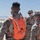A group of military personnel in desert camouflage uniforms are gathered in an outdoor arid environment, appearing to conduct a briefing or coordination session. Several individuals are wearing bright orange safety vests with reflective strips, with one vest marked '43T'. The setting appears to be a training exercise at a desert military installation.