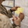 A military personnel member in desert camouflage (MARPAT) uniform is writing on a yellow index card or note card with a pink marker while standing in front of a tactical map board. The map in the background appears to show a geographic area with roads, boundaries, and place names including 'Emerson Lake' and 'Acorn'. The scene suggests a military planning or operations center environment.