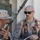 Two military personnel in desert camouflage (MARPAT) uniforms are engaged in communication during what appears to be a training exercise or operational activity. One individual is speaking into a handheld radio while the other, wearing dark sunglasses and an orange-accented harness vest with multiple attached items, listens attentively. The setting appears to be an outdoor military installation or training area.