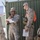 Two U.S. Marines in MARPAT desert camouflage uniforms are standing near a green military shipping container. The Marine on the left is holding a radio (appears to be an AN/PRC-152 or similar tactical radio) and reviewing a document, while the Marine on the right carries multiple radios, an orange backpack, and holds a spiral notebook. The setting appears to be a forward operating base or training environment.