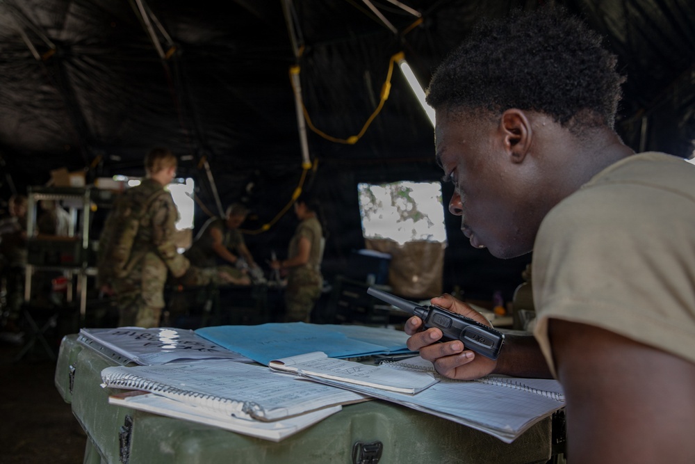 A military personnel in a tan uniform is leaning over a field desk inside a tactical tent, holding a handheld radio while reviewing documents and notebooks. The setting appears to be a forward command post or operations center during a field exercise or deployment. Several other uniformed soldiers are visible in the background engaged in various tasks.