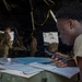 A military personnel in a tan uniform is leaning over a field desk inside a tactical tent, holding a handheld radio while reviewing documents and notebooks. The setting appears to be a forward command post or operations center during a field exercise or deployment. Several other uniformed soldiers are visible in the background engaged in various tasks.