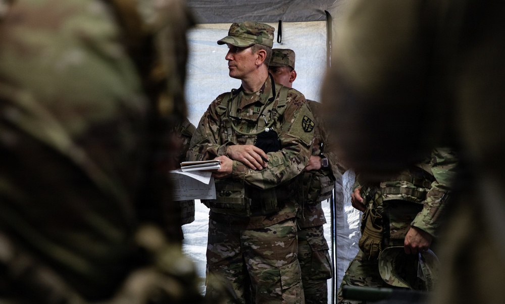 A group of military personnel in MultiCam/OCP camouflage uniforms are gathered inside a tent or temporary shelter structure. The central figure, appearing to be a senior officer or NCO, is holding documents and appears to be conducting or attending a briefing. The scene suggests a field operations or training environment.