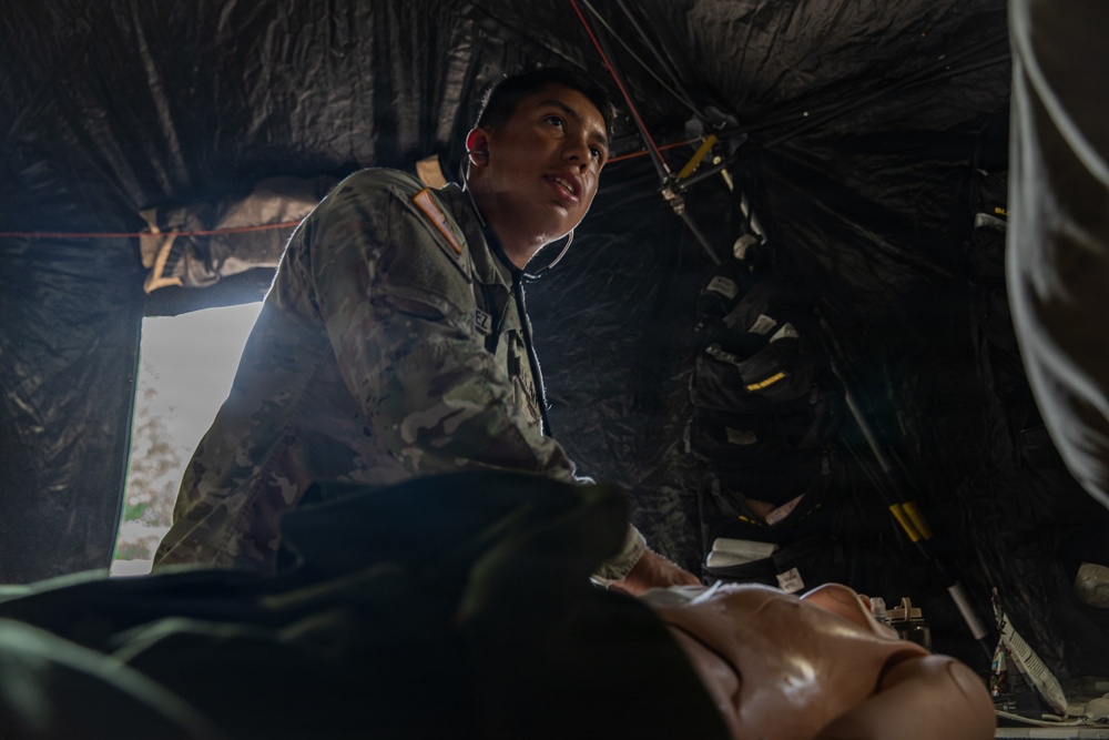 A soldier in military camouflage uniform wearing a stethoscope is attending to a patient or training mannequin inside what appears to be a field medical tent. The low-angle perspective shows the medic leaning over the subject, suggesting active medical assessment or treatment. The setting appears to be a military field exercise or forward operating medical station.