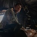 A soldier in military camouflage uniform wearing a stethoscope is attending to a patient or training mannequin inside what appears to be a field medical tent. The low-angle perspective shows the medic leaning over the subject, suggesting active medical assessment or treatment. The setting appears to be a military field exercise or forward operating medical station.