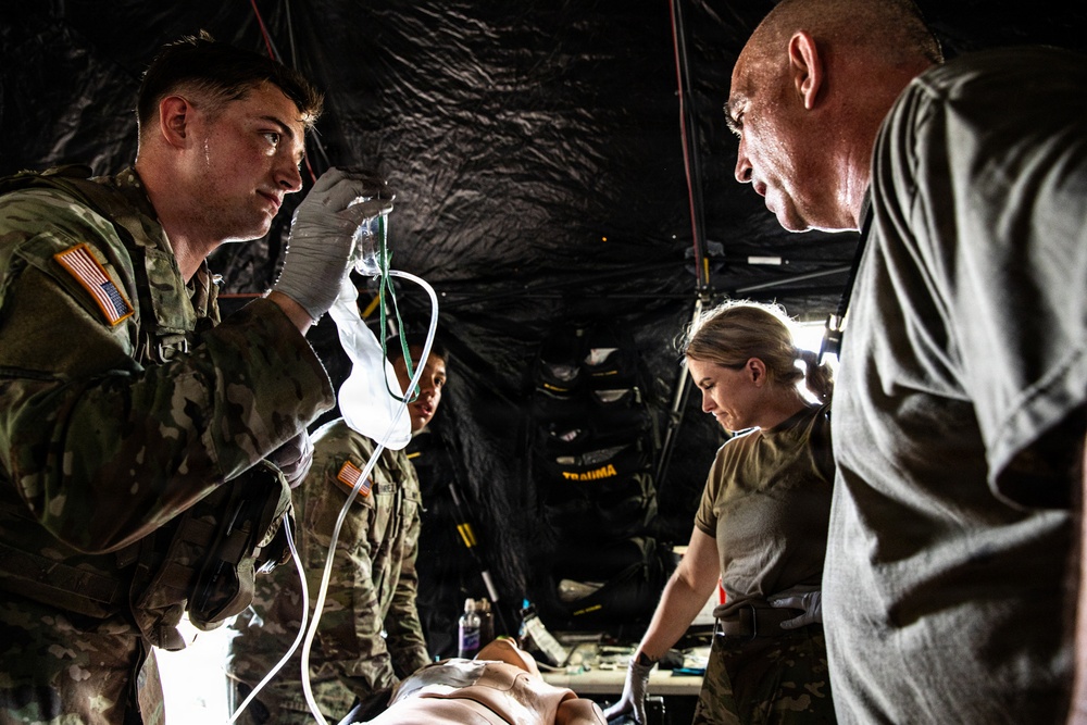 Military medical personnel are conducting what appears to be a field medical training exercise inside a tactical tent or shelter. Soldiers in U.S. Army camouflage uniforms are working on a medical simulation mannequin, with one soldier holding IV tubing and another performing hands-on procedures. A civilian or senior instructor in a gray shirt observes and appears to be evaluating or directing the trainees.