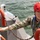 CTF 66 Conducts Pier-Side Testing and Control of K3 Scout Craft A military personnel member wearing US Navy digital camouflage (NWU Type III) and a red NRS helmet is working on or near a watercraft. The individual is wearing a blue life vest and holding onto an orange-painted railing with a chain attached. The setting appears to be a maritime environment with open water visible in the background.