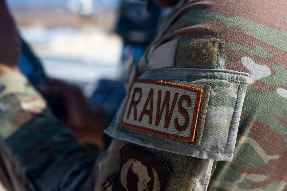 Close-up photograph of a military uniform sleeve showing a 'RAWS' patch on OCP (Operational Camouflage Pattern) multicam fabric. The patch features dark red lettering on a tan/khaki background with an orange border. A second partially visible patch appears below it, and a velcro field is visible above.