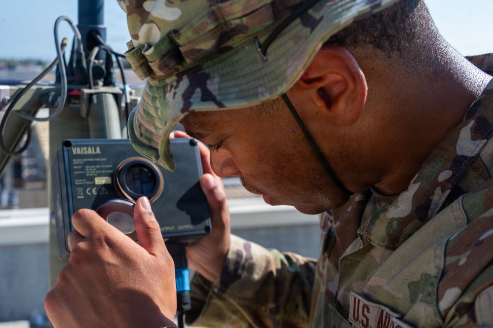 A U.S. Army soldier in OCP (Operational Camouflage Pattern) uniform is closely inspecting or working on a Vaisala brand meteorological or sensor device. The soldier is leaning in with focused attention, appearing to examine or adjust a lens or port on the equipment. The setting appears to be an outdoor rooftop or elevated platform.
