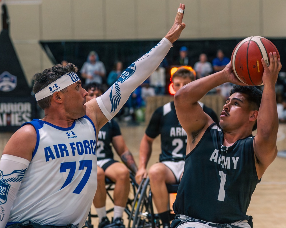 A wheelchair basketball game is in progress between U.S. Air Force and U.S. Army teams, likely at the Warrior Games. An Army player wearing jersey number 1 is attempting a shot while an Air Force player wearing number 77 reaches up to contest the shot. Spectators are visible in the background watching the competitive adaptive sports event.