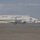 A twin-engine turboprop aircraft, consistent with a Beechcraft King Air or similar military variant, is taxiing on a wet tarmac at a military or civilian airfield. A large white hangar dominates the background along with what appears to be a control tower structure to the right. The image has a hazy, overcast quality likely due to recent rainfall and atmospheric moisture.
