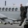 A military personnel member in an olive green flight suit is descending the airstairs of a United States Marine Corps aircraft on a tarmac. The gray twin-engine turboprop aircraft bears the text 'UNITED STATES MARINE' on its fuselage. The setting appears to be a military airfield with additional aircraft visible in the background.