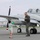 A military aviator in olive green flight suit laughs and reacts playfully as a young boy aims a colorful water gun at him on an airport tarmac. The scene appears to be a joyful reunion or welcome-back moment in front of a twin-engine turboprop aircraft marked with 'UNITED S...' on the fuselage. The interaction conveys warmth and humor between what appears to be a servicemember and a child, likely a family member.