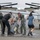 A group of people on a military airfield tarmac are dousing a person in military flight suit with buckets of water in what appears to be a celebratory tradition, likely a 'fini flight' or retirement ceremony. Multiple individuals are participating, including civilians and military personnel, with at least two large buckets being emptied simultaneously. A military tiltrotor aircraft, consistent with a V-22 Osprey, is visible in the background.