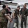 A group of military personnel, appearing to be U.S. Marines based on their camouflage uniforms, are participating in a traditional 'fini flight' or retirement/milestone water dousing ceremony on an airfield tarmac. One individual in a flight suit is being soaked with water from a large bucket while others laugh and participate enthusiastically. The celebratory nature and aviation setting suggest this is a milestone aviation tradition.